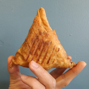A Hand Holds Up A Samosa Against A Plain Gray Background.