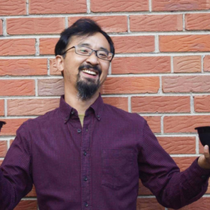 A headshot of Howard. He’s wearing a dark purple button-up shirt and stands against a brick wall. He has dark hair, a cropped goatee, and wears glasses.