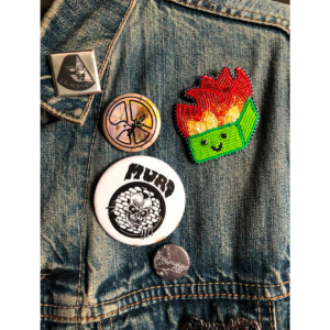 A Close-up Photo Of A Jean Jacket Featuring Several Pins And Patches. One Is A Beadworked Dumpster Fire.