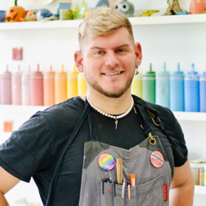 A Photo Of A Person In An Apron, They Stand In Front Of A Wall Displaying A Rainbow Array Of Paint In Squeeze Bottles.
