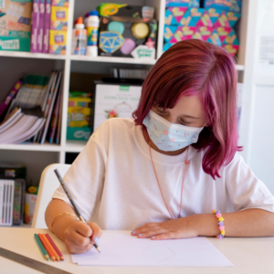 A Child With Pink Hair Sits At A Desk, They Are Working With Some Pencil Crayons.