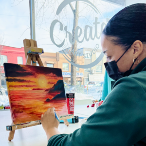 A Person Sits At The Create Art Studio Window Working On A Small Painting Of A Sunset.