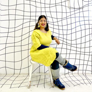 A portrait of Khadija Aziz. She sits on a white stool against a backdrop of wavy grid of black lines. She's wearing a bright yellow dress, yellow earrings and is smiling at the camera.