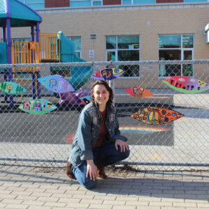 Sylvie Crouches In Front Of A Chain Link Fence Decorated With Painted Wooden Cutouts. They Are Hand-painted In A Childish Style Featuring Different Scenes, One Is Of A Soccer Field, Another Is Of A Tree House In A Forest.