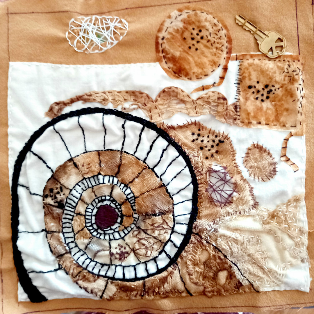 A tea-stained swatch of fabric with a spiral pattern embroidered into it. Various other types of fabric and black and white stitches form an abstract collage around the spiral and as a part of the spiral.