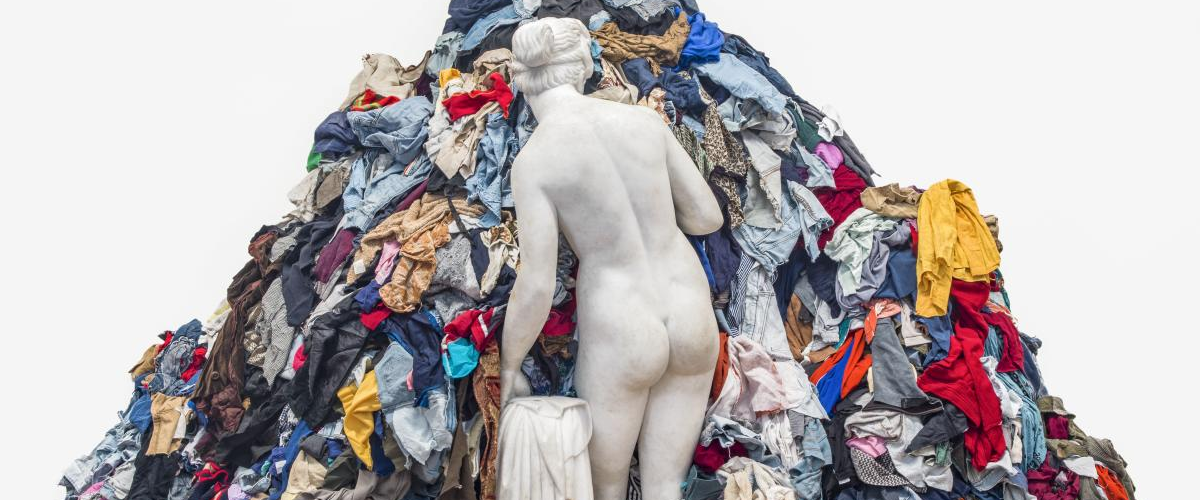 A white marble statue of a female figure carrying a cloth faces into a massive pile of discarded clothing.