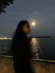 A photo of a person standing by the water under the full moon's light.