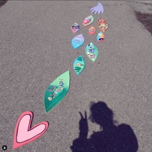 A Series Of Colourful Wooden Cutouts Depicting Various Scenes In A Naive Art Style. They Are Laid On The Ground, In The Corner, Sylvie’s Shadow Throws A Peace Sign.